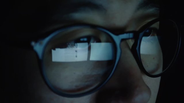 Asian young man scrolling the website in the computer with reflection on glasses in dark bedroom - the website being used is public domain