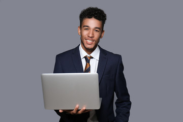 Handsome African-American businessman with laptop on grey background