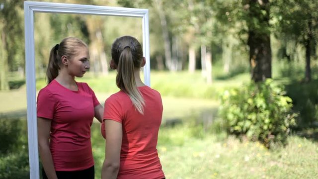 Two twin girls in sports clothes in the park as a reflection in the mirror. scene with a mirror image