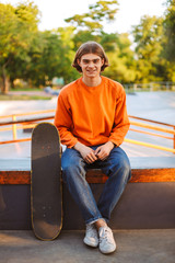 Obraz premium Young smiling skater in orange pullover and jeans happily looking in camera with skateboard at skatepark