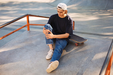 Young skater in cap holding his painful leg with skateboard near while spending time at skatepark