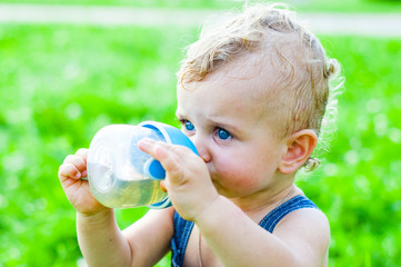little baby boy sitting on the grass in a park and drinking from the bottle.