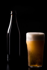 Glass of crafted unfiltered beer with foam and bottle behind isolated on black dark background with soft reflections and beautiful shadows. Studio shot.