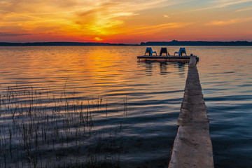 Fototapeta na wymiar Three empty chairs on wooden jetty on lake, during sunrise.