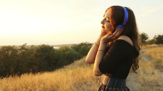teenager girl in headphones listening to music on nature, young woman walking in the field at sunset