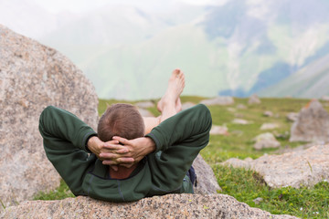 a man with hands behind his head is lying on a stone in highland relaxing and looking into the distance at the mountains