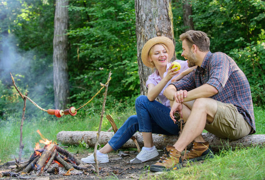 Couple relaxing sit on log having snacks. Family enjoy romantic weekend in nature. Girl offers eat apple while they waiting roasted food. Picnic roasting food over fire. Have snack. Cooking at picnic