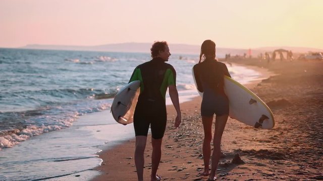 SLOW MOTION: A young couple of guy and girl surfers are walking along the beach in wetsuits. In the hands of holding surfboards. Look at the waves and the sunset