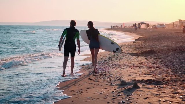 SLOW MOTION: A young couple of guy and girl surfers are walking along the beach in wetsuits. In the hands of holding surfboards. Look at the waves and the sunset