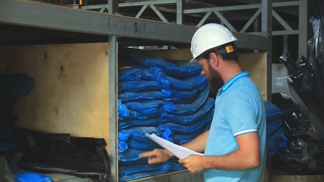 a worker in a white helmet in the warehouse checks the availability of the goods on the invoice.