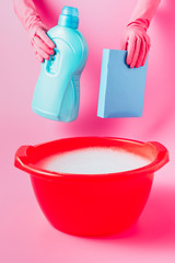 cropped image of female cleaner in rubber gloves holding laundry liquid and washing powder over plastic basin with foam, pink background