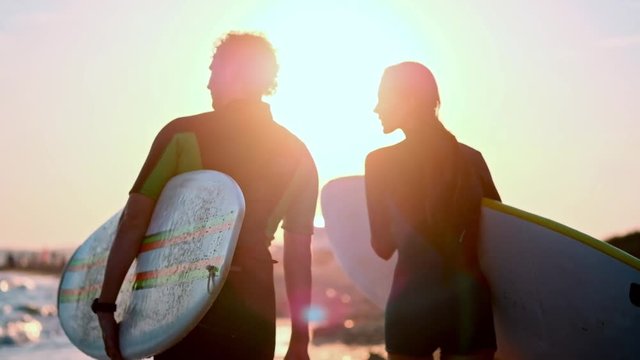 SLOW MOTION: A young couple of guy and girl surfers are walking along the beach in wetsuits. In the hands of holding surfboards. Look at the waves and the sunset