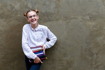 Portrait of a cute smiling schoolgirl 12 years old with glasses, with books. Gray concrete background wall in school with copyspace. Funny girl