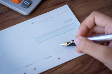 Female hand writing blank cheque book with calculator on the wooden table background.