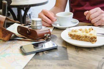 A man is holding a smartphone. He is sitting at the cafe table on the street. On the table, there is a vintage camera, a tourist map, a coffee and a piece of cake on a plate.