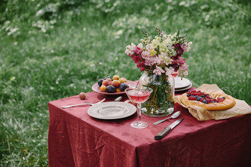 wineglass, berries pie and bouquet of flowers on table in green garden