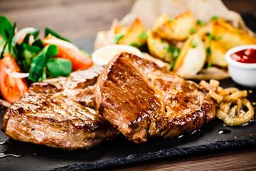 Grilled steaks with baked potatoes and vegetables served on black stone on wooden table 