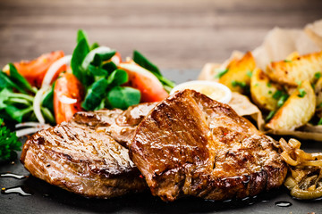 Grilled steaks with baked potatoes and vegetables served on black stone on wooden table 