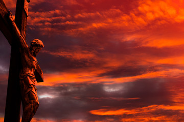Jesus on a cross against burning red sunset sky with clouds and little patches of light coming...