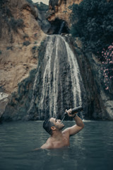 Young man drinking a cold beer. Man enjoying a beer and bathing in a waterfall.