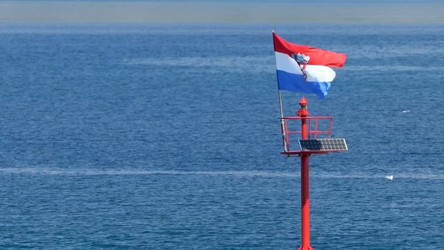 Stone pier with lighthouse and flying Croatian flag in daylight, steady