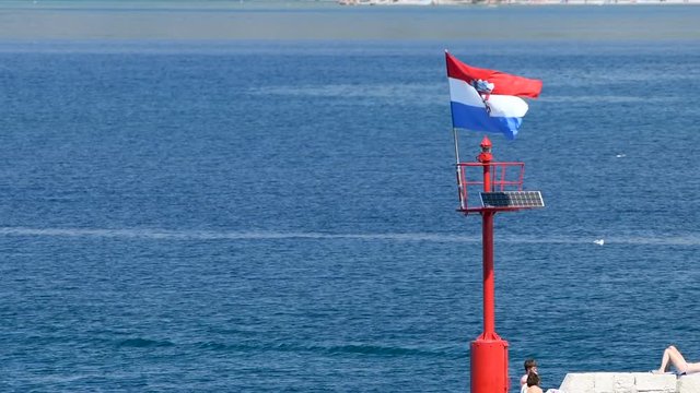 Stone pier with lighthouse and flying Croatian flag in daylight, people walking and sitting on stone wall, , zoom out