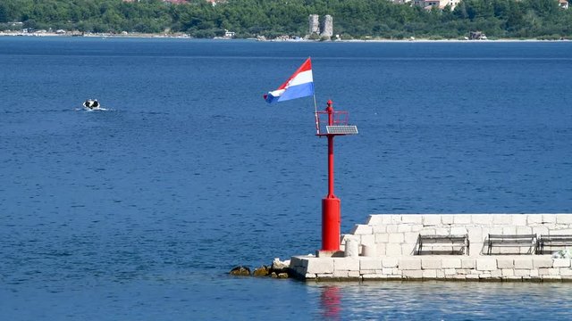 Stone pier with lighthouse and flying Croatian flag in daylight, motor boat in background, people unrecognizable
