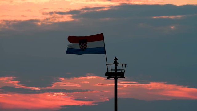 Stone pier with lighthouse and flying Croatian flag in sunset