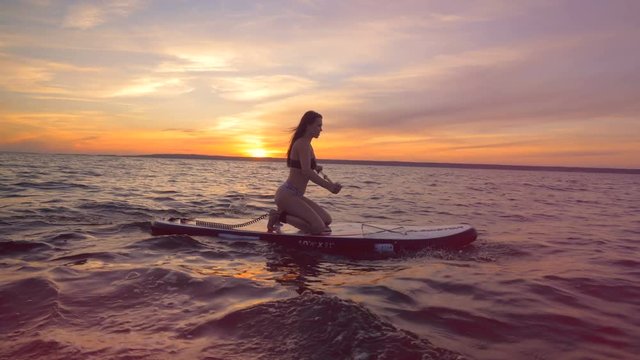 A woman rows on a sunset background, side view. A girl rides her surfing board, going in the ocean.
