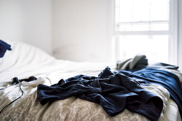 Messy room with dark blue black clothes lying on top of bed sheets comforter in depressing cold...