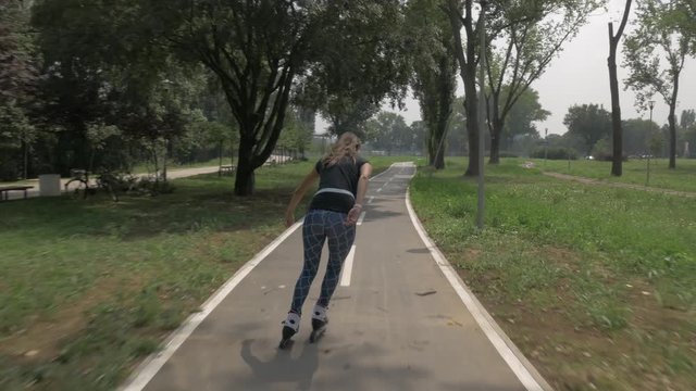 White handsom brown hair girl in tight blue pants woman riding roller skates on bike road in park, during summer day