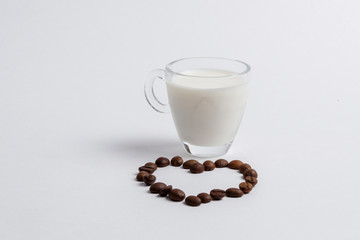 A glass mug full of milk is standing next to coffee beans in the form of a heart on a white background