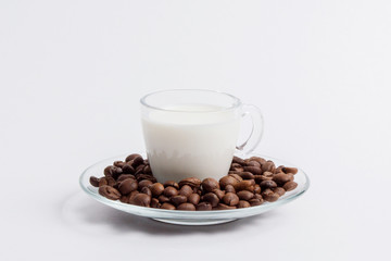 A glass mug full of milk is standing in a pile of coffee beans on a saucer on a white background