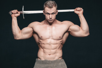Bodybuilder man posing with a sword isolated on black background. Serious shirtless man demonstrating his mascular body