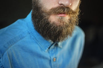 Attributes of the Appearance of a Hipster Male: Mustache and Beard Close Up. Portrait of Bearded Man in a Blue Denim Shirt on Black Background