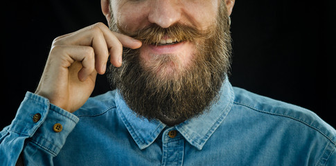 Smiling Bearded Man in a Blue Denim Shirt Twists the Mustache with His Hand on a Black Background....
