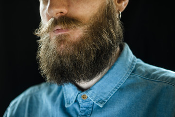Fototapeta premium Attributes of the Appearance of a Hipster Male: Mustache and Beard Close Up. Portrait of Bearded Man in a Blue Denim Shirt on Black Background