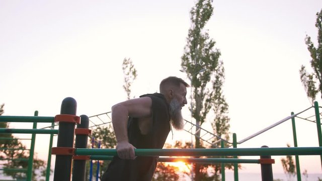 Fit handsome middle aged man with long gray beard doing push ups on parallel bars on sports ground with sea background