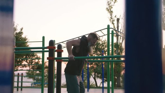Fit handsome middle aged man with long gray beard doing push ups on parallel bars on sports ground with sea background