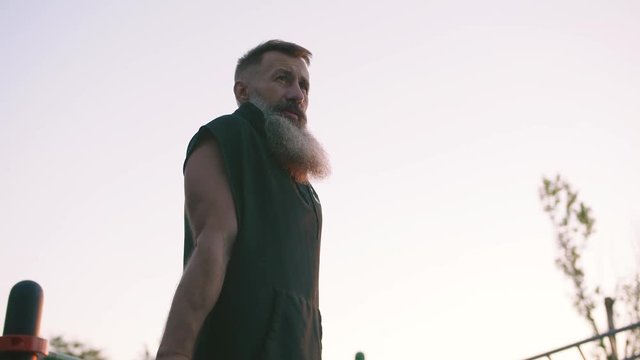 Fit handsome middle aged man with long gray beard doing push ups on parallel bars on sports ground with sea background