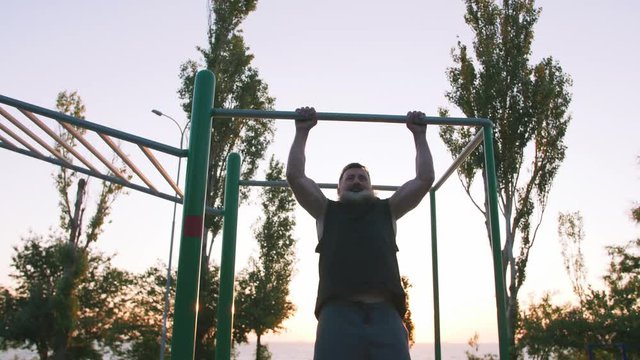Fit handsome middle aged man with long gray beard doing pull-ups on horizon bar on sports ground with sea background