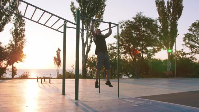 Fit handsome middle aged man with long gray beard doing pull-ups on horizon bar on sports ground with sea background