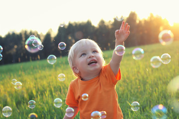 Cute toddler blond boy playing with soap bubbles on summer field. Happy child summertime concept. Authentic lifestyle image - obrazy, fototapety, plakaty