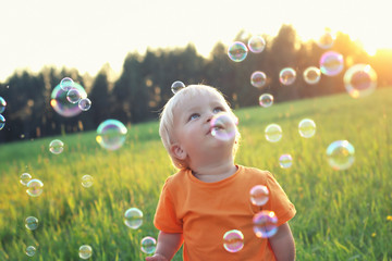 Cute toddler blond boy playing with soap bubbles on summer field. One bubble on his head. Happy childhood concept. Authentic lifestyle image