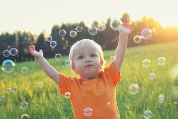 Cute toddler blond boy playing with soap bubbles on summer field. Hands up. Happy childhood...