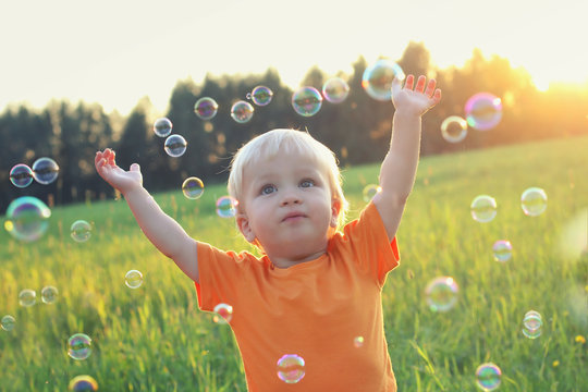 Cute toddler blond boy playing with soap bubbles on summer field. Hands up. Happy childhood concept. Authentic lifestyle image