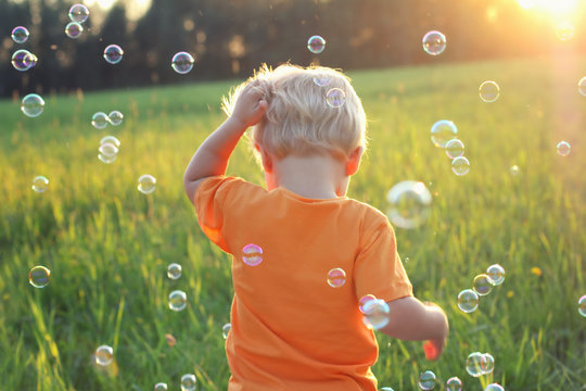 Cute toddler blond boy playing with soap bubbles on summer field. He is standing with his back. Authentic lifestyle image