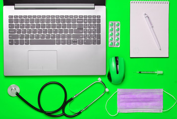 Workplace of a modern doctor. Laptop, wireless mouse, stethoscope, pills, syringe on green pastel background. Top view, minimalism.
