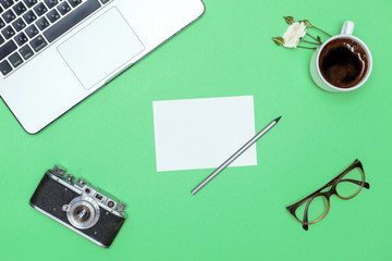 office desk with a laptop a cup of coffee, glasses, a blank and camera on a bright green background with space for text. Top view with copy space, flat lay
