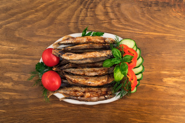 Small fish fried in oil on a porcelain plate with red whole tomatoes, slices of pepper and cucumber, dill sprigs, parsley and basil  leaves.